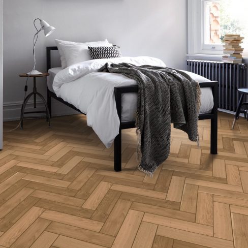 A close-up of HE405 herringbone wood flooring, showcasing precisely cut planks arranged in a classic herringbone pattern. The surface features a natural wood grain texture with warm, neutral tones, enhancing the sophisticated and timeless aesthetic.