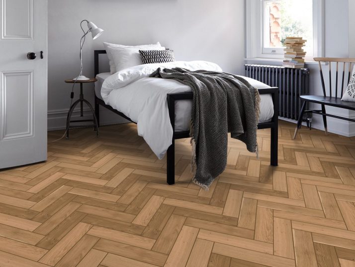 A close-up of HE405 herringbone wood flooring, showcasing precisely cut planks arranged in a classic herringbone pattern. The surface features a natural wood grain texture with warm, neutral tones, enhancing the sophisticated and timeless aesthetic.